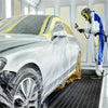 A person in a protective suit is applying pre-taped masking film to a silver car for painting protection.