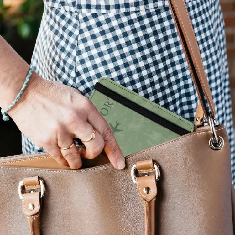 A hand places an RFID blocking leather card holder and passport wallet into a tan leather handbag.