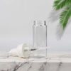 A clear glass bottle and a white nozzle are shown on a marble surface with a green palm leaf in the background.