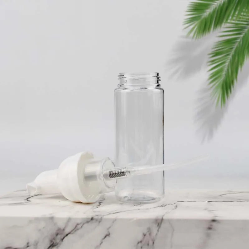A clear glass bottle and a white nozzle are shown on a marble surface with a green palm leaf in the background.
