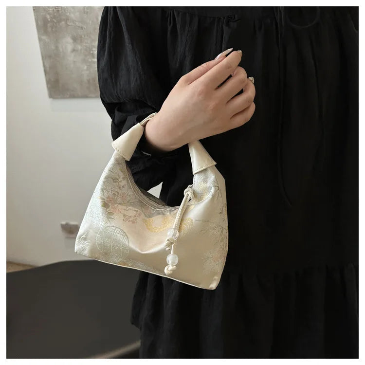Nahaufnahme einer cremefarbenen Seiden-Crossbodytasche mit Perlenstickerei, gehalten von einer Person in einem schwarzen Klei
