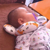 A baby is resting on a brown surface, supported by a white pillow with orange designs and a purple safety strap.