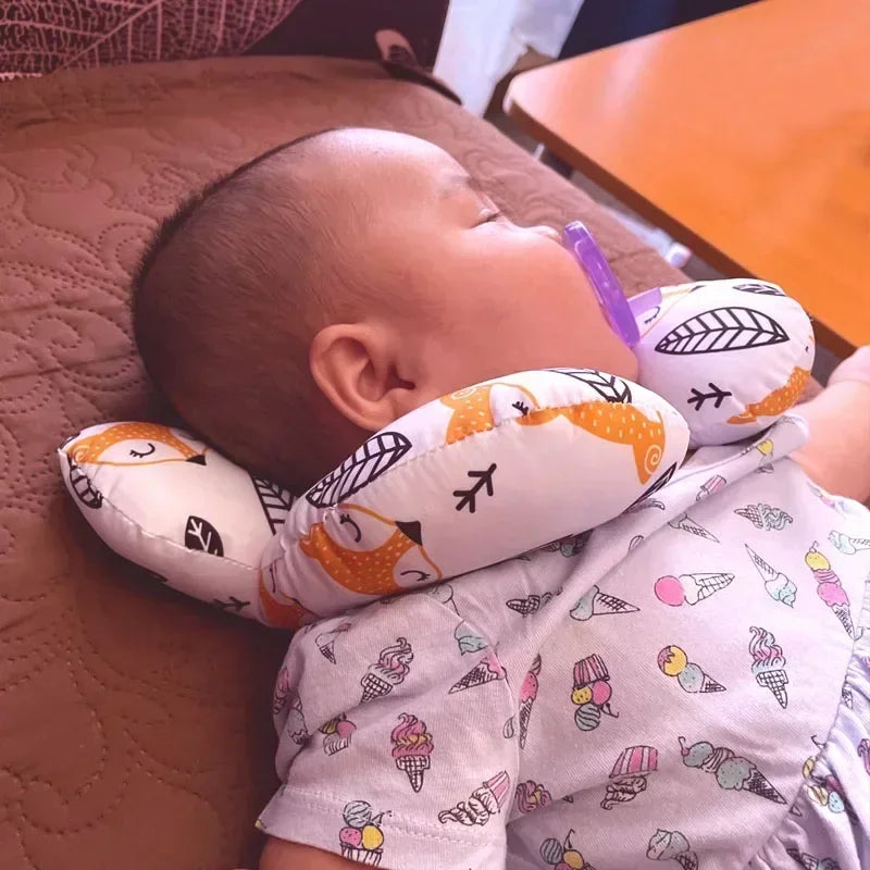 A baby is resting on a brown surface, supported by a white pillow with orange designs and a purple safety strap.