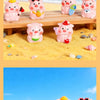 A collection of pink resin pig figurines in various poses and colors displayed on a wooden surface amidst yellow sand.