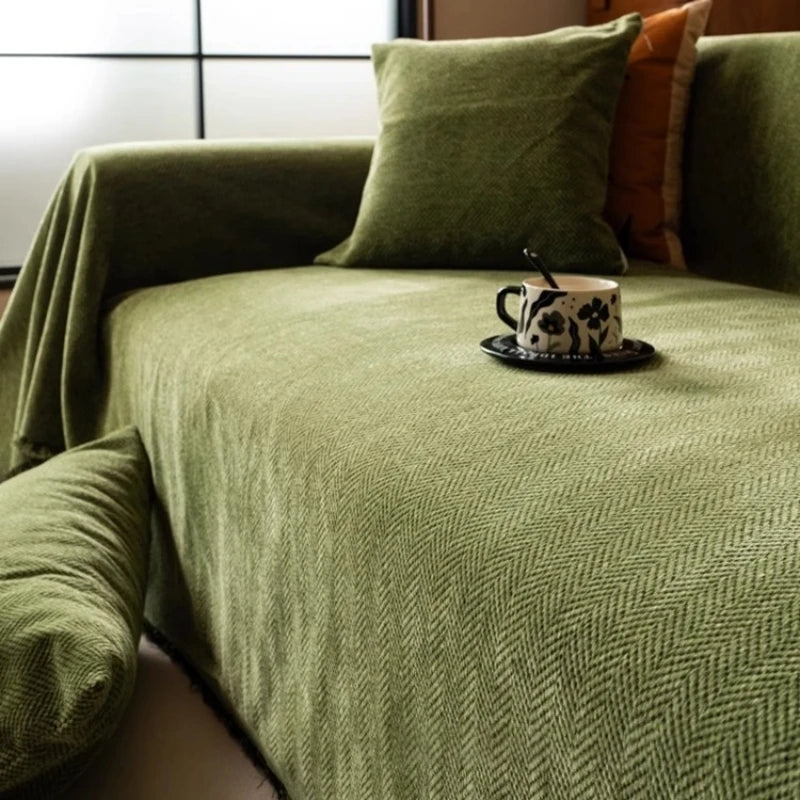 A close-up of a moss green herringbone chenille sofa throw draped over a sofa with orange accent pillows.