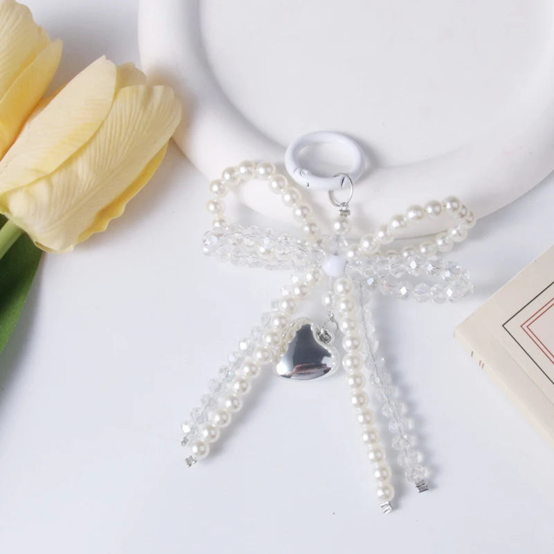 A clear pearl bow keychain with a heart charm resting on a white ceramic object next to a yellow tulip.