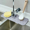 A silicone kitchen sink mat with a faucet splash guard and a wooden brush are displayed on a stainless steel sink.