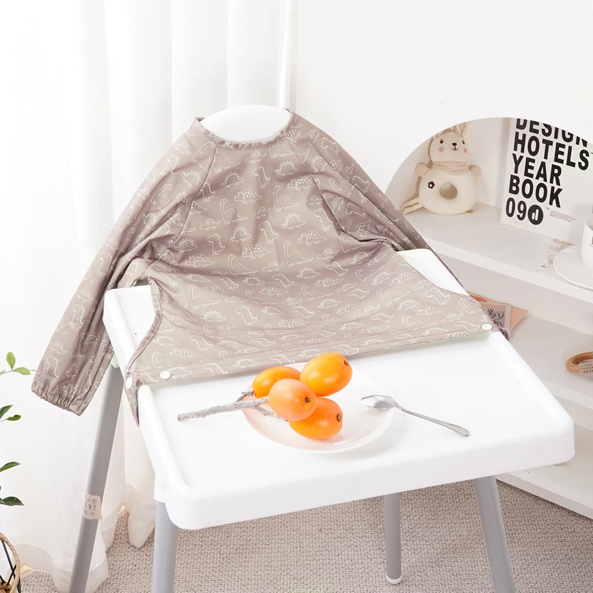 A beige long-sleeve waterproof baby bib with a cute animal design displayed on a white table.