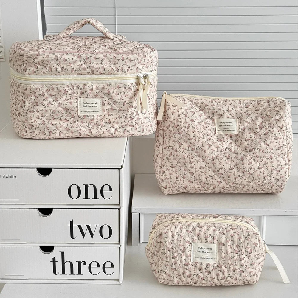 A set of three vintage floral quilted cosmetic bags in light pink and cream colors displayed on a white shelf.