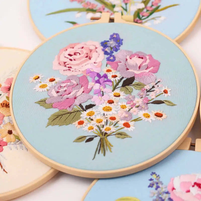 A close-up of a floral embroidery starter kit featuring pink, purple, and white flowers on a light blue fabric.