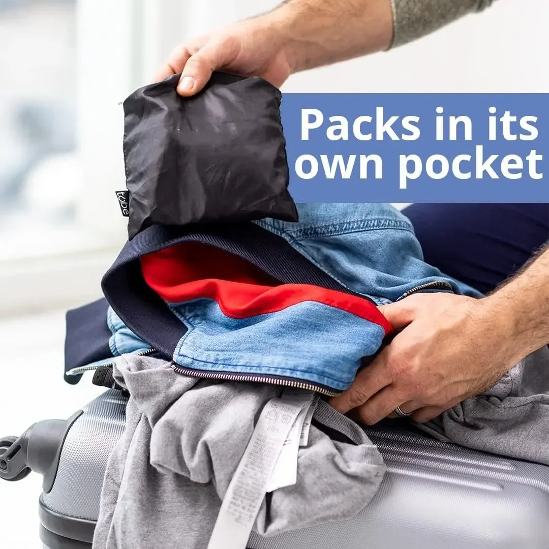 A hand is placing a black fabric pack into a pocket of a blue denim jacket, demonstrating the self-filling travel pillowcase 