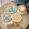 A set of colorful floral diamond art coasters on a woven mat next to a cup of latte and a gold spoon.