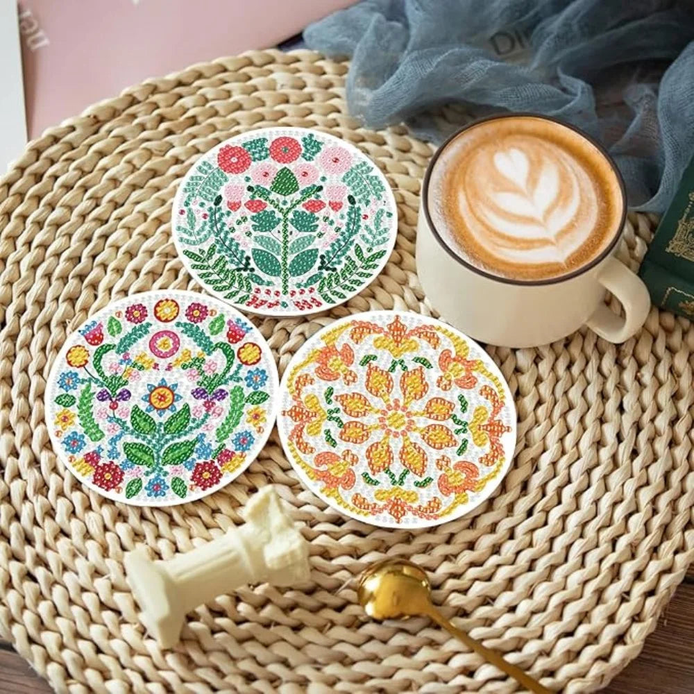 A set of colorful floral diamond art coasters on a woven mat next to a cup of latte and a gold spoon.