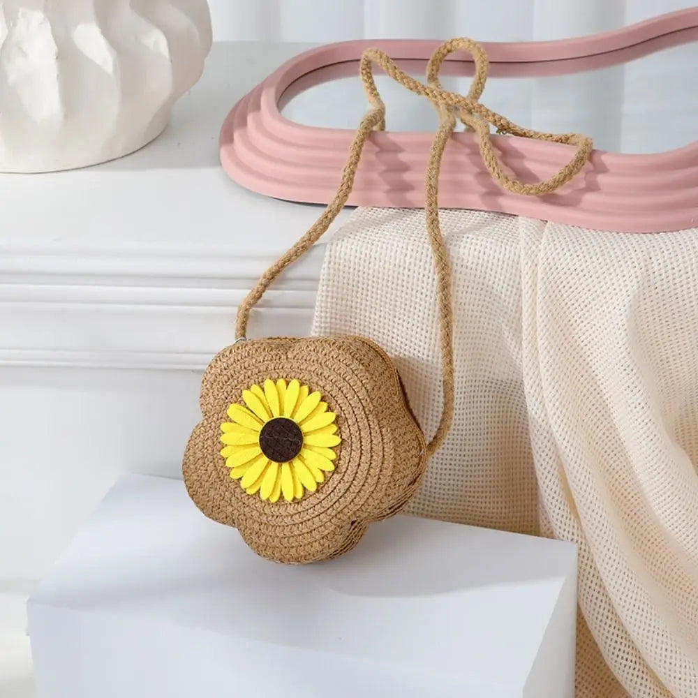 Sac bandoulière en paille tissée en forme de tournesol sur une surface blanche, à côté d'un plateau rose et d'un tissu drapé.