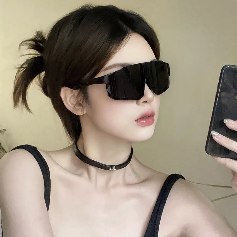 A close-up of a woman wearing black cycling sunglasses and a black choker, looking towards the camera.