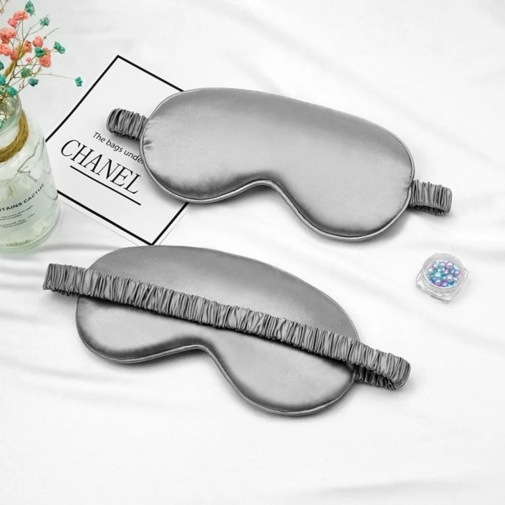 A gray silk sleep mask with a fabric strap is displayed on a white background next to a box labeled CHANEL.