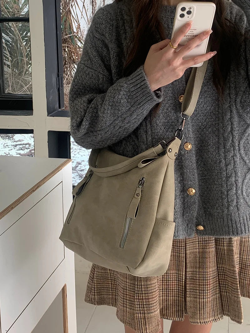 A woman holds a beige matte leather crossbody hobo bag with an adjustable strap against a light background.