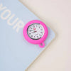 A pink silicone pocket watch with a brooch pin is shown resting on a light blue notebook.