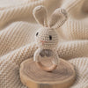 A handmade crochet bunny toy with long ears and a natural wood teether ring resting on a beige knitted fabric.