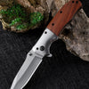 A folding pocket knife with a walnut wood handle and stainless steel blade rests on a dark surface next to rocks.