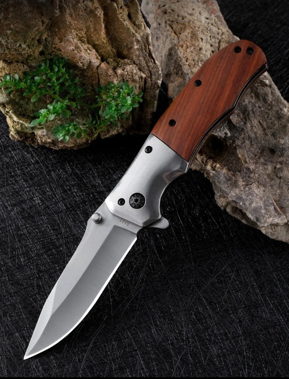 A folding pocket knife with a walnut wood handle and stainless steel blade rests on a dark surface next to rocks.