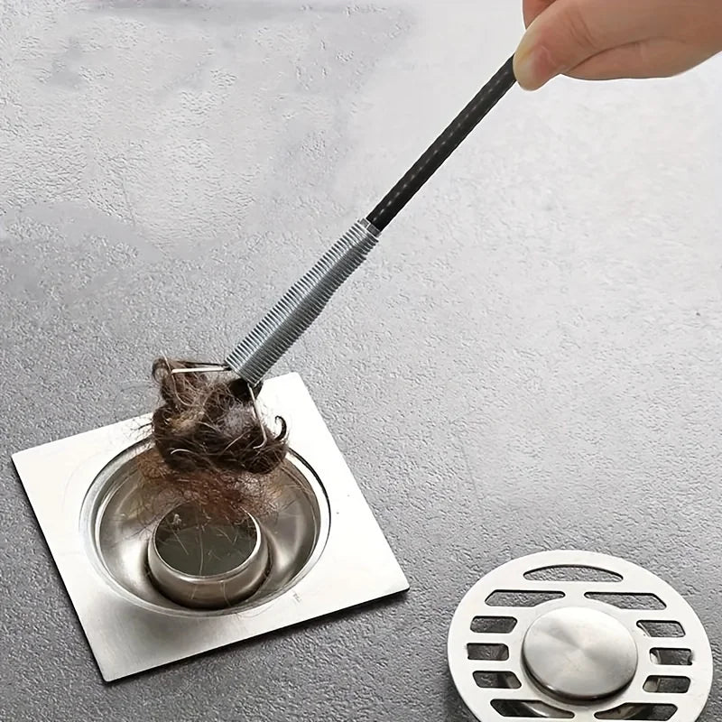 A hand is using a black drain auger with a claw grabber to remove hair from a drain.