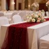 A deep red pearl beaded tulle table runner draped on a white tablecloth for an elegant wedding or event setting.