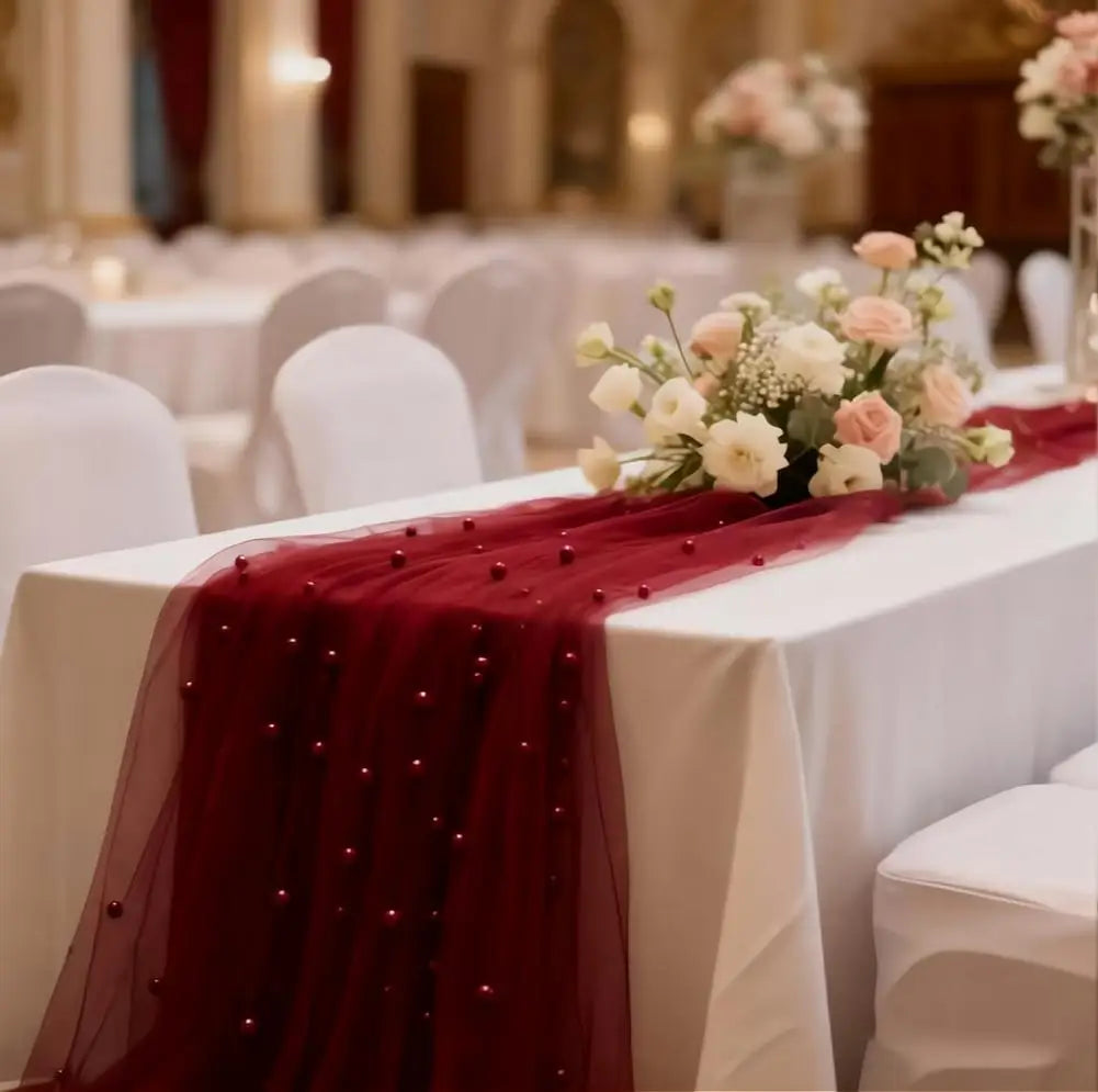 A deep red pearl beaded tulle table runner draped on a white tablecloth for an elegant wedding or event setting.