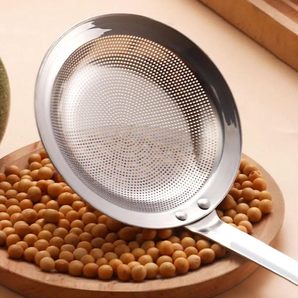 A stainless steel fine mesh skimmer spoon is shown resting on a wooden board surrounded by small, round, light brown objects.