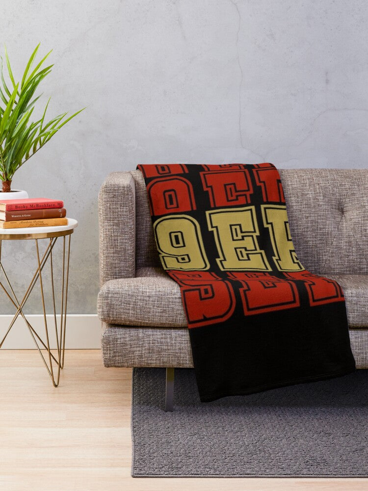 A knit throw blanket featuring the NFL team logo in red, gold, and black on a gray sofa.