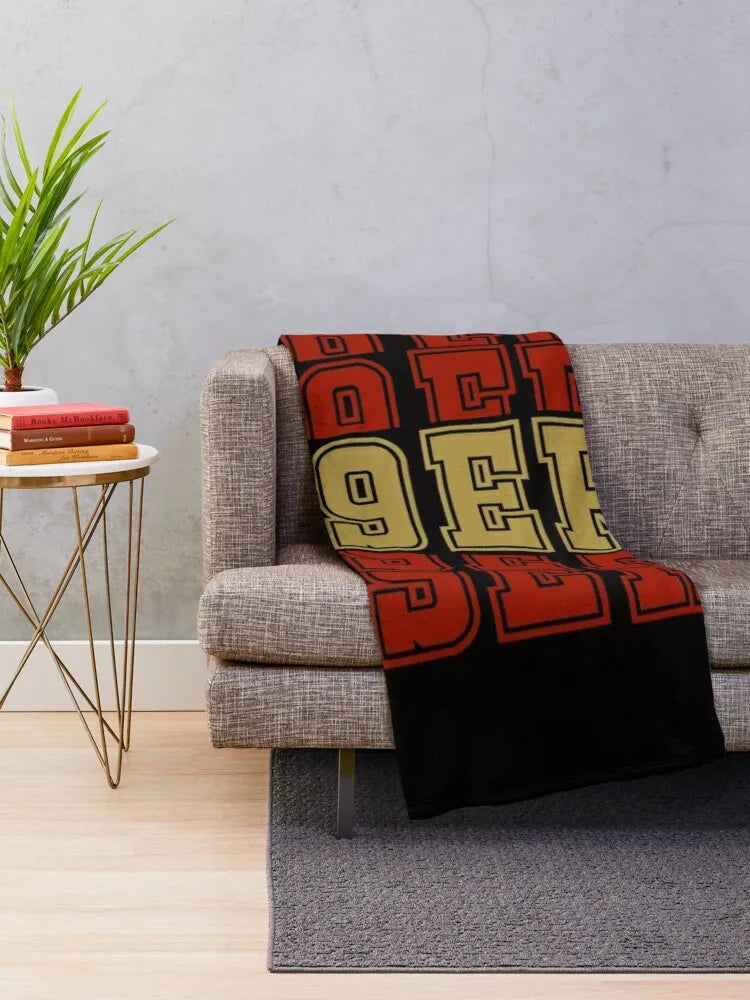 A knit throw blanket featuring the NFL 49ers logo in red, gold, and black on a gray sofa.