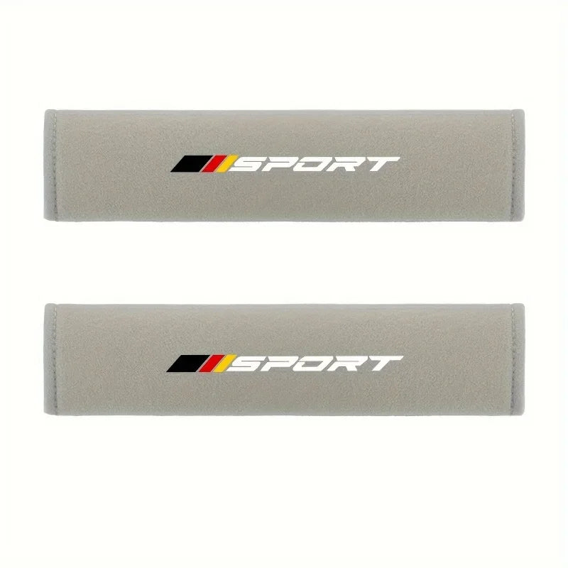 A pair of beige soft car seat belt cushions featuring the Sport logo in black and orange.