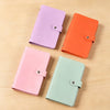 Four colored cash organizer binders in purple, orange, pink, and mint green with snap closures on a wooden surface.