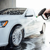 A hand sprays water onto a white car with a pressure washer gun to clean the exterior.