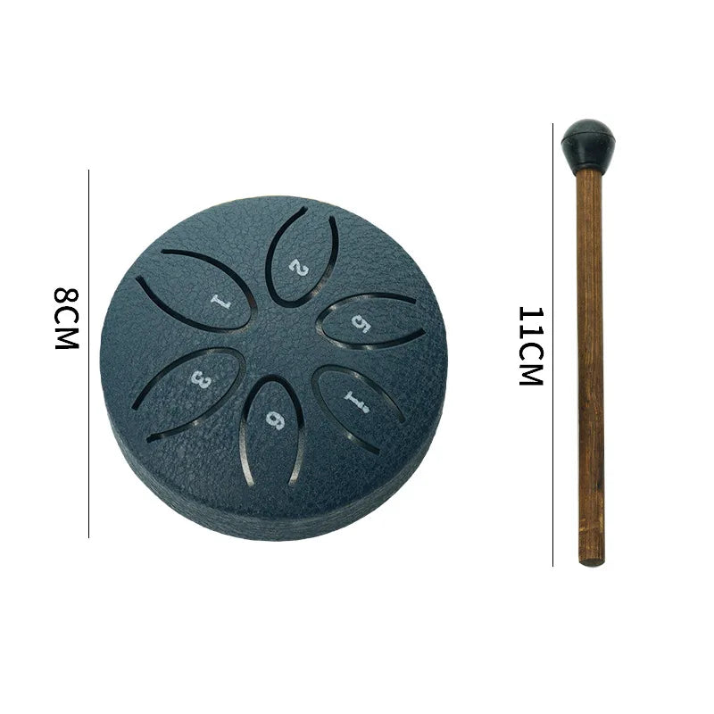 A dark gray mini steel tongue drum with six tuning holes and a wooden mallet shown next to a measurement scale.
