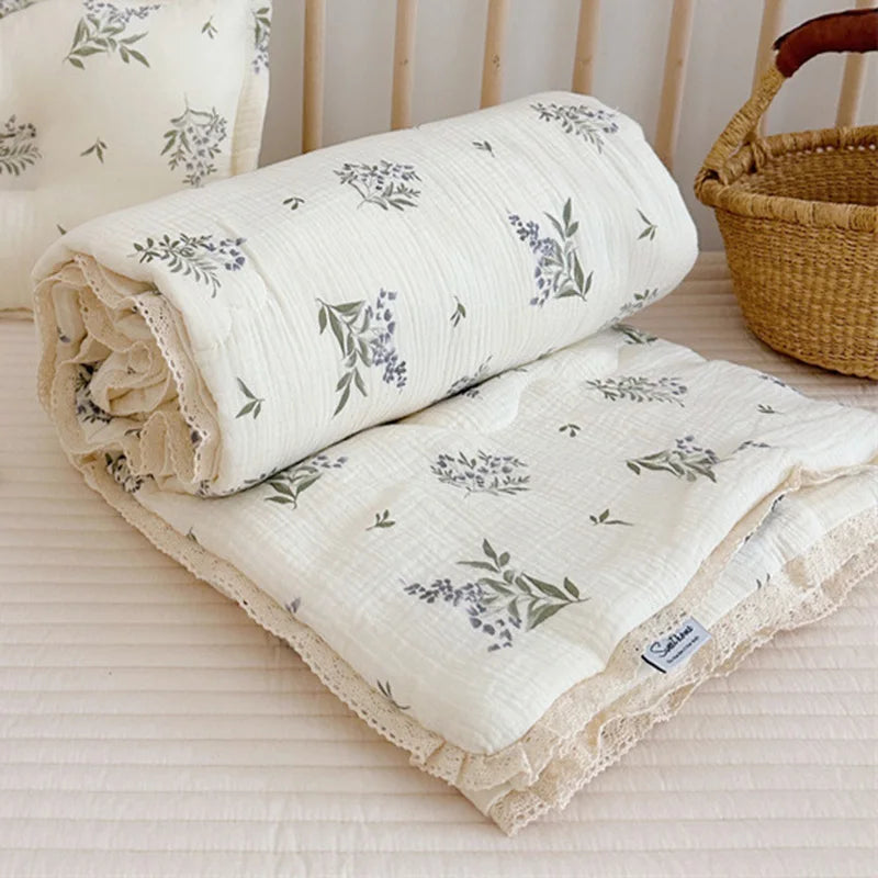 A rolled white floral muslin baby blanket with gray and green leaves resting on a light pink surface.