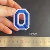 A hand holds a blue embroidered letter Q patch, measuring 5.3 cm high.