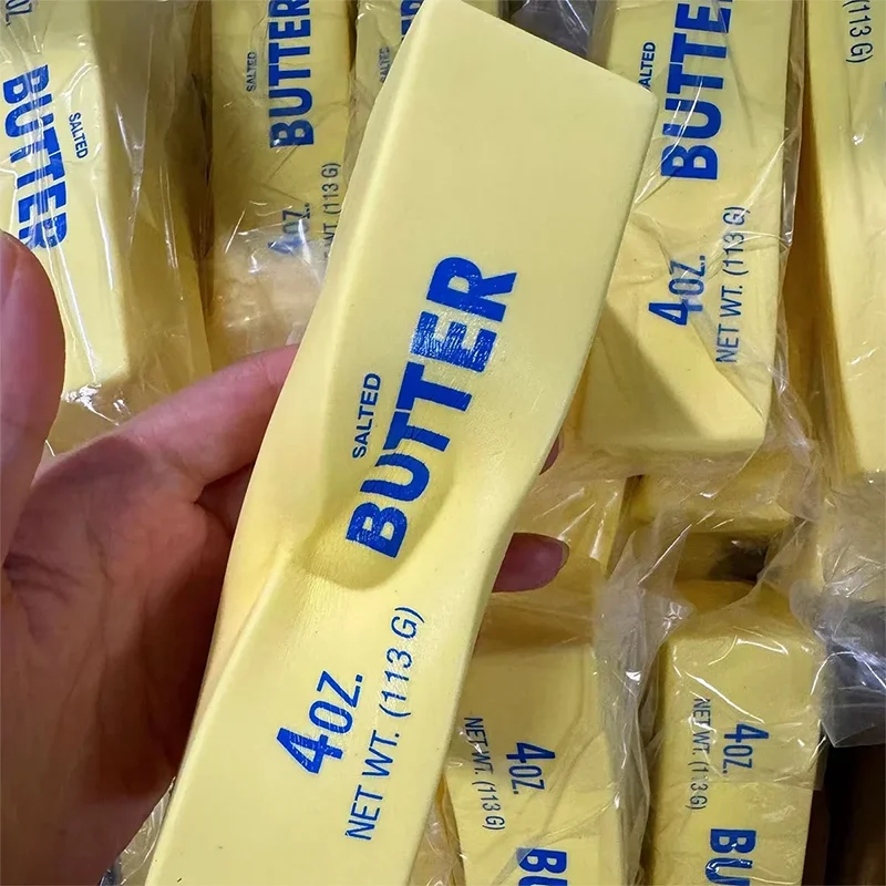 A hand holds a yellow butter stick fidget toy with the word BUTTER printed on it, packaged in clear plastic.