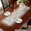 A lace table runner with a floral pattern is displayed on a wooden table set for an elegant meal.