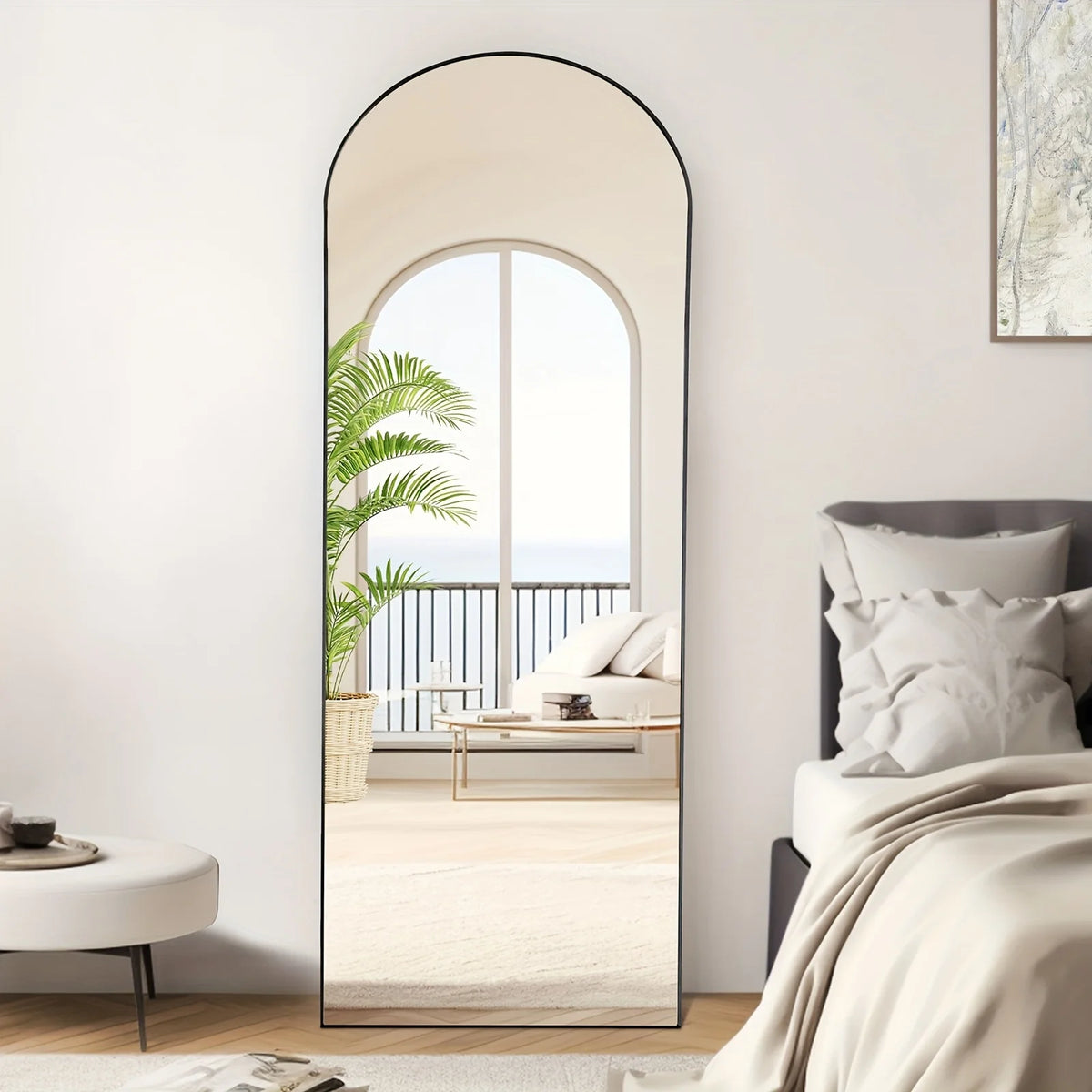 An arched full-length floor mirror with an aluminum frame reflecting a bright interior and outdoor view.