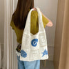 A woman is holding a white handwoven knit tote bag with blue swirl and star patterns against a light background.