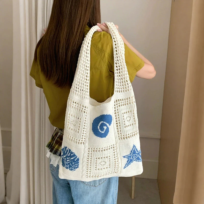 A woman is holding a white handwoven knit tote bag with blue swirl and star patterns against a light background.