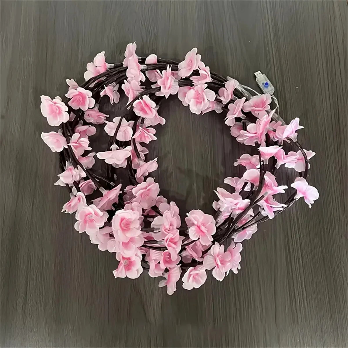 A circular wreath made of pink cherry blossom flowers strung on LED lights with a USB power connector.