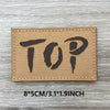 A brown woven patch with the word TOP printed in black text on a gray wooden surface.