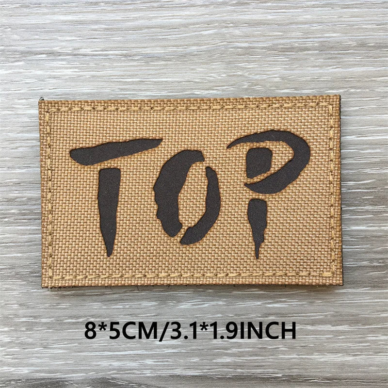 A brown woven patch with the word TOP printed in black text on a gray wooden surface.