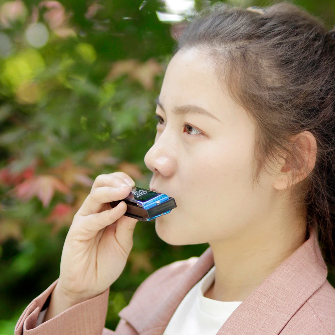 A young woman holds a blue harmonica in its protective case against a blurred green outdoor background.