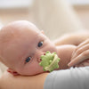 A close-up of a baby's face being gently touched by a silicone baby teether toy shaped like a flower and grape.