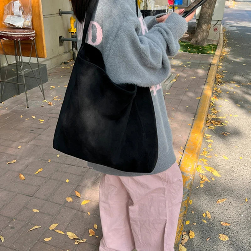 A person wearing a gray hoodie and pink pants is carrying a black suede tote shoulder bag outdoors.