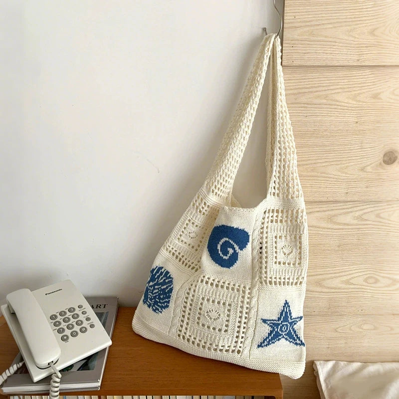 A cream-colored handwoven knit tote bag with blue swirl and star patterns resting on a wooden surface.