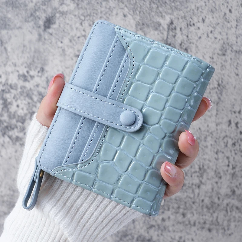 A light mint green crocodile-embossed clutch wallet is held in a hand against a gray background.